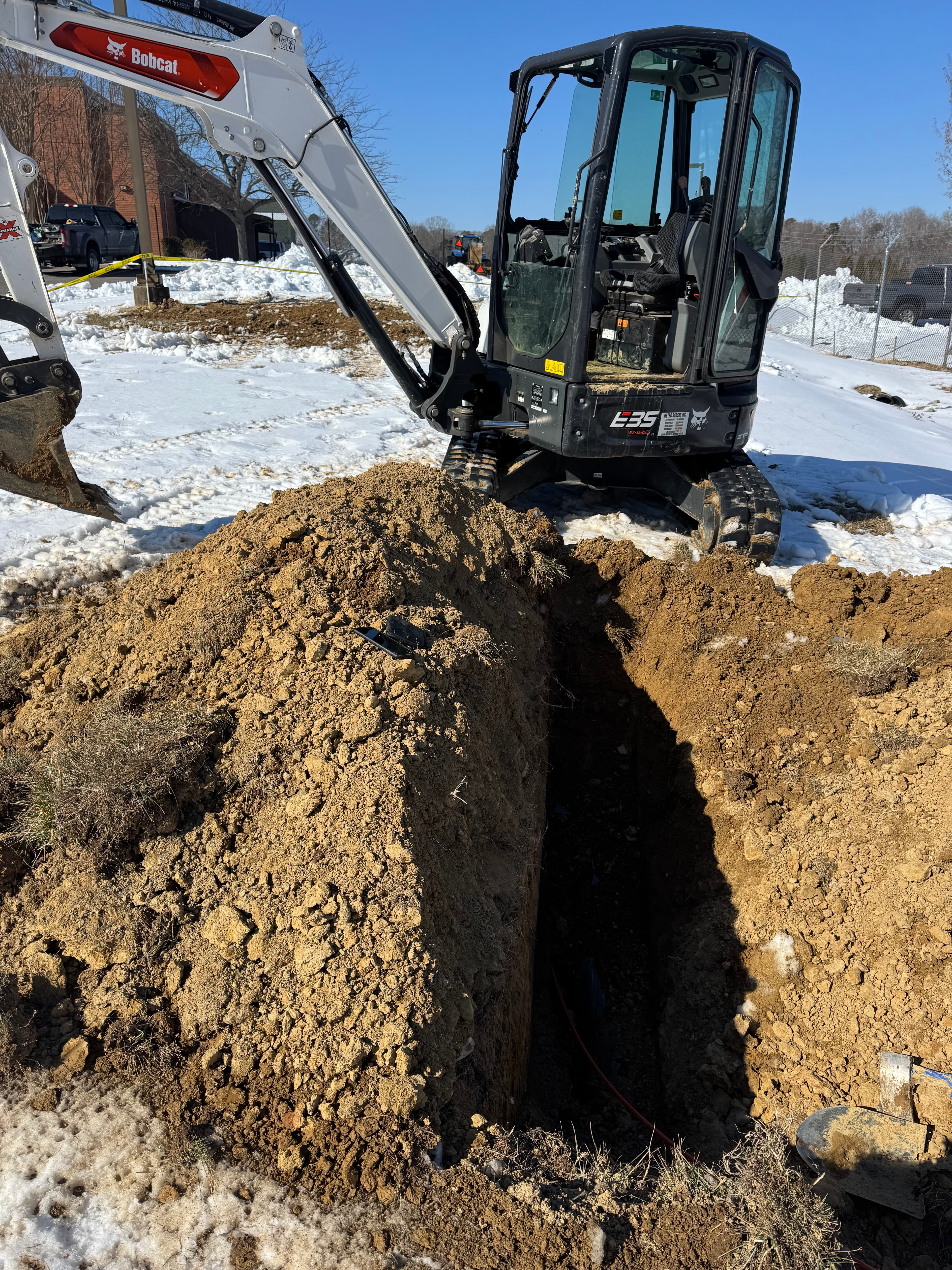 Bobcat excavator parked beside a deep trench in a snowy field under a clear blue sky.