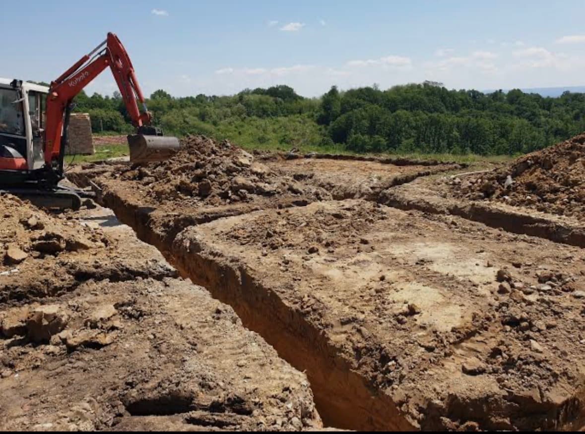 Orange excavator digging deep foundation trenches in a dirt field overlooking a lush green forest.