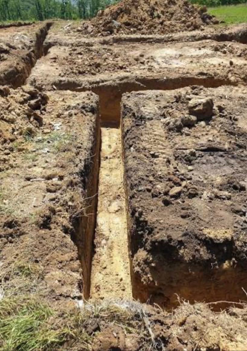 Deep rectangular trenches excavated in brown soil at an outdoor construction site with dirt piles nearby.