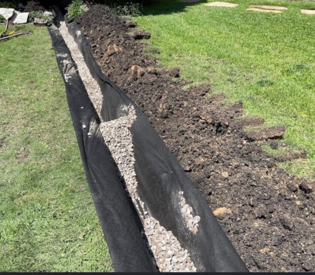 French drain trench lined with black fabric and filled with gravel in a grassy yard.