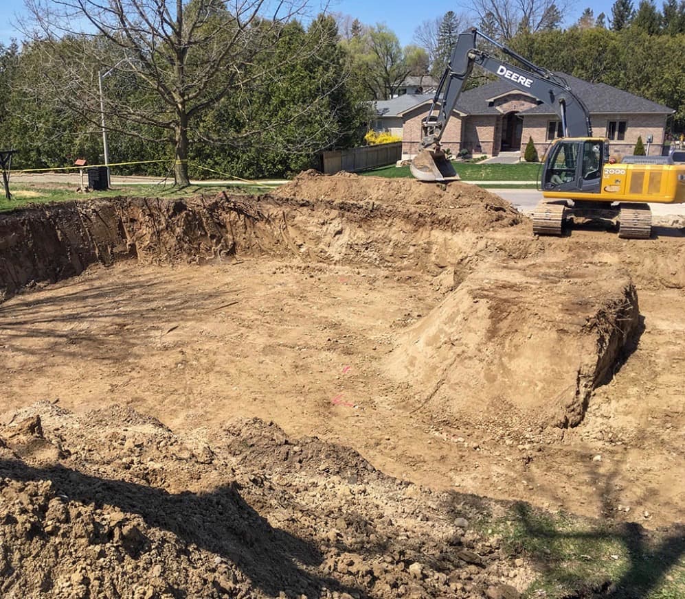 Yellow John Deere excavator parked beside a large dirt pit in a residential backyard.