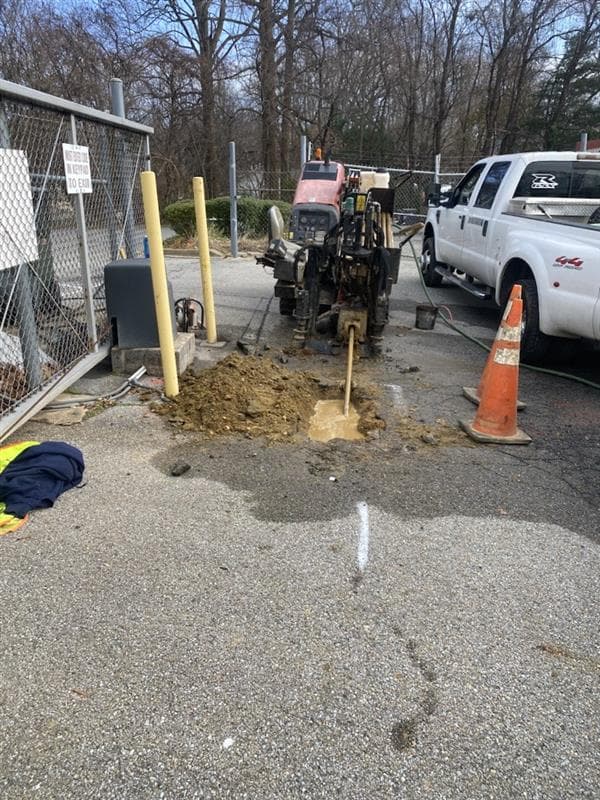 An orange directional drill rig bores into the ground in an asphalt parking lot.