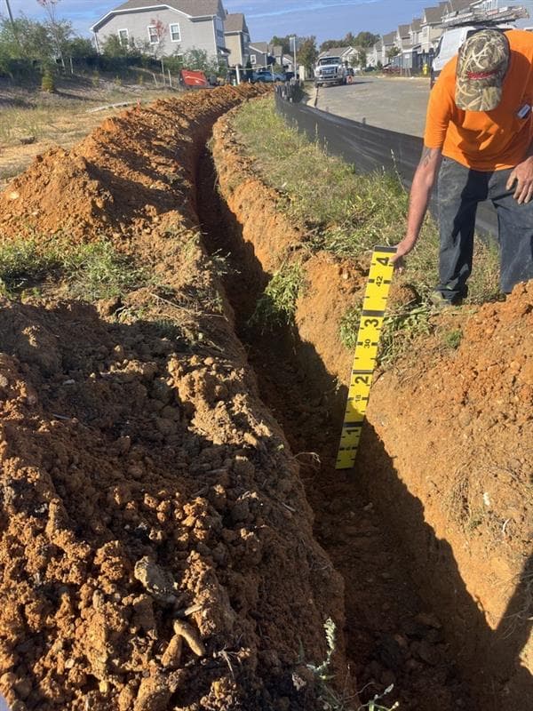 Worker measuring the depth of a long dirt trench using a yellow measuring stick.