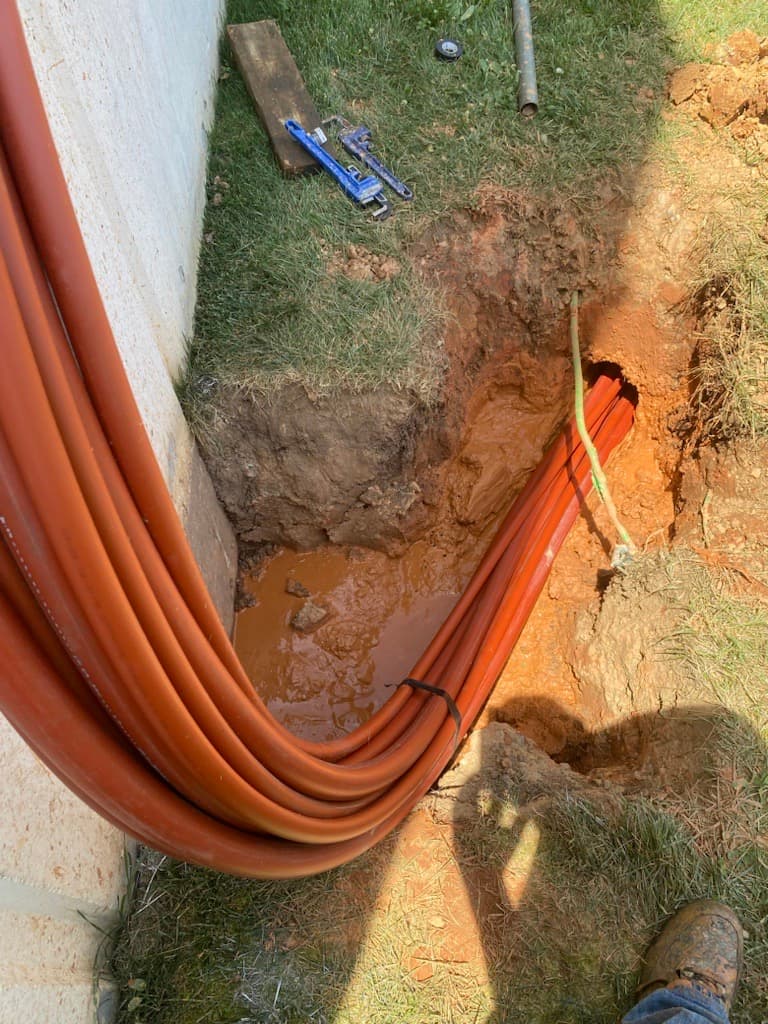 Bundle of orange pipes curving into a muddy trench next to a concrete foundation wall.