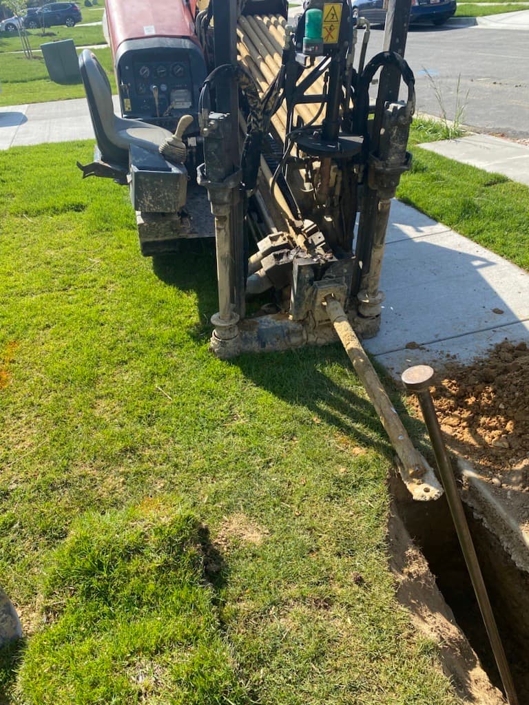Horizontal directional drilling machine installing underground pipes in a residential lawn next to a sidewalk.