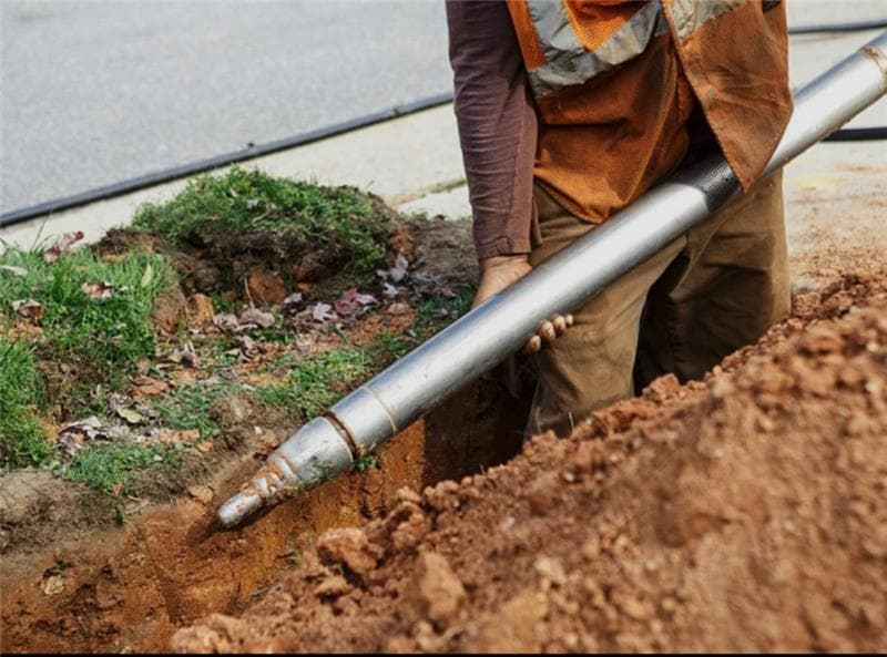 Construction worker guides a large metal piercing tool into a trench for underground utility installation.