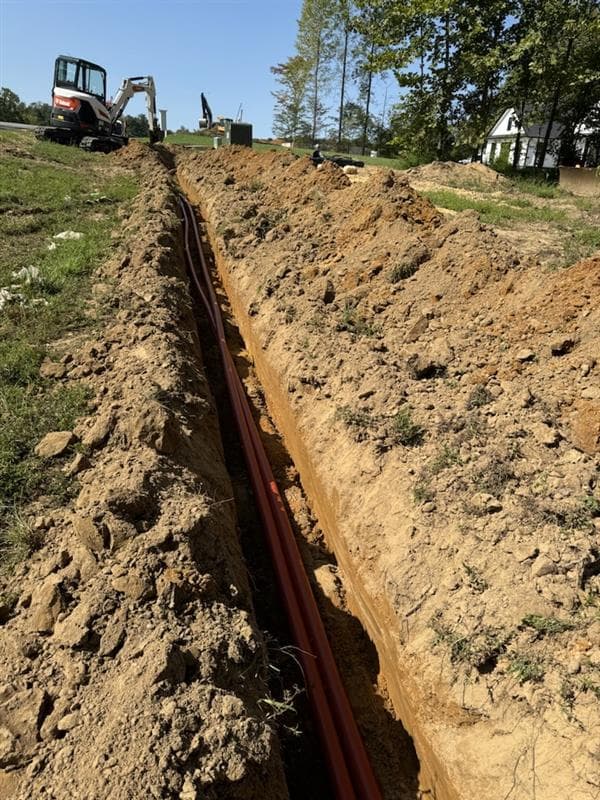 Red utility pipes laid in a long dirt trench with a white excavator nearby.