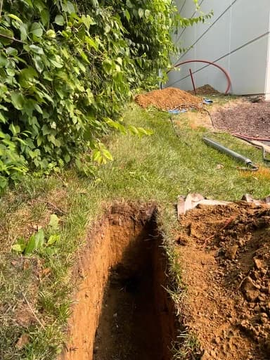 Narrow trench dug into a grassy lawn beside a pile of excavated brown dirt.