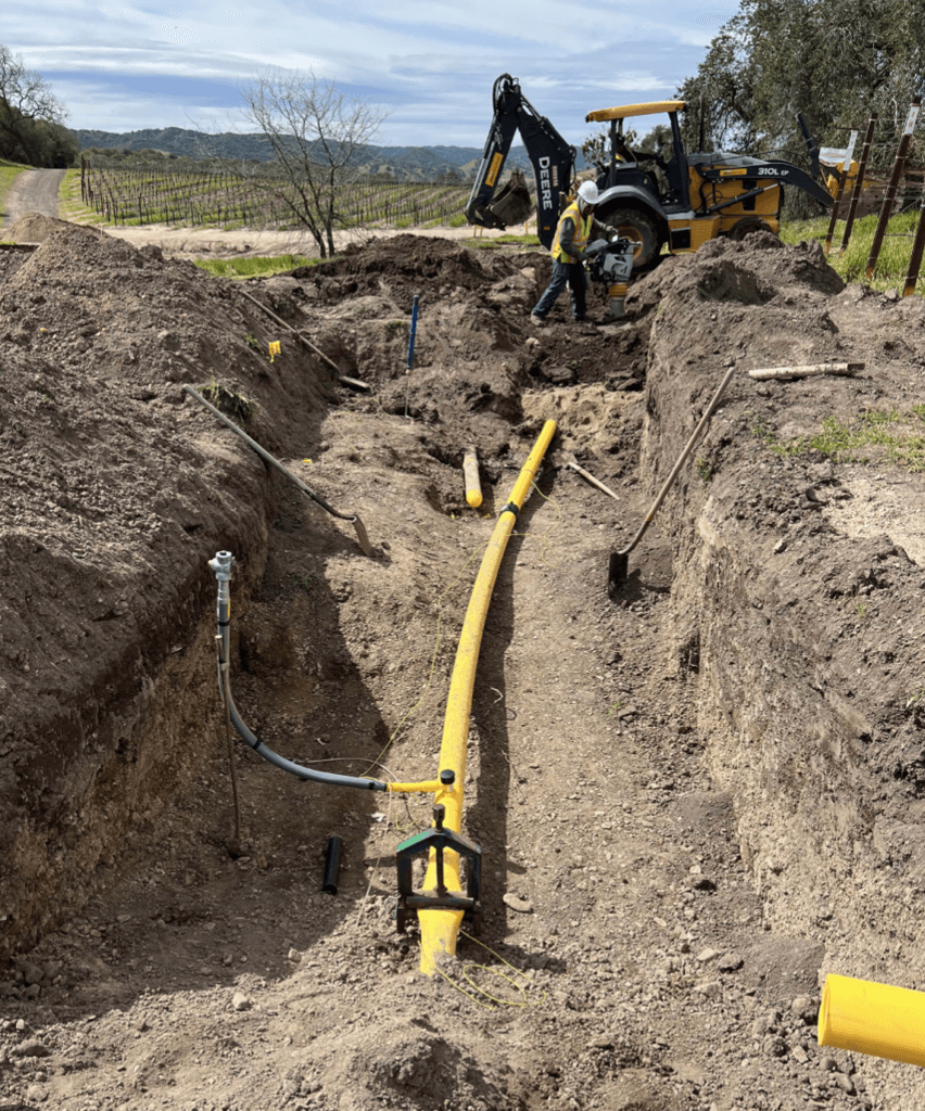 Construction worker installs a yellow pipe in a dirt trench with a backhoe nearby.
